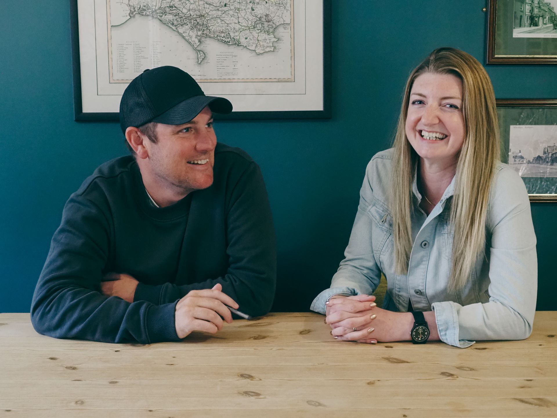 Two people sit at a wooden table smiling, with a teal wall behind them displaying a historical map of Italy and framed photographs.